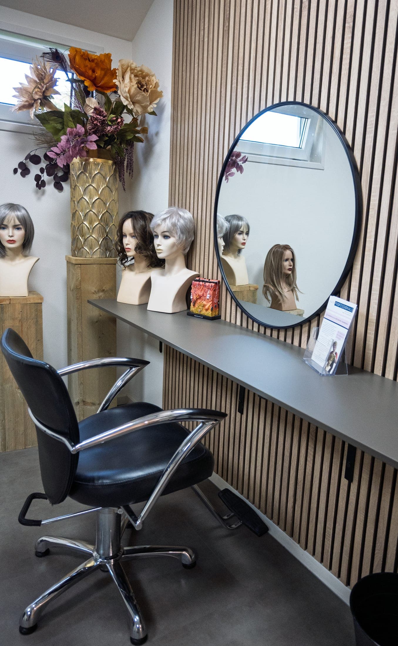 Een behandelplek bij Hair Professionals met een stoel voor de spiegel en verschillende pruiken op standaards.